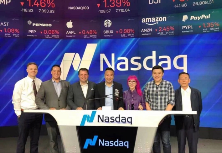 Seven individuals pose, smiling, behind the Nasdaq desk at the New York Stock Exchange. At the center are Xin Wu and Xiaoyan Huang. Huang, the only woman in the group, has bright pink hair.